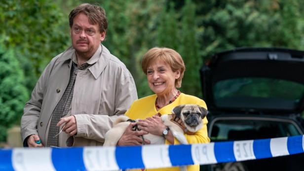 Katharina Thalbach als Miss Merkel hält vor Absperrband Mops in Arm