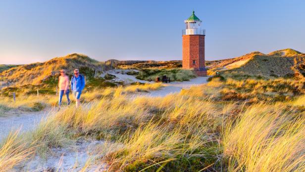 Zwei Personen gehen auf einem sandigen Weg durch Dünen, im Hintergrund steht ein roter Leuchtturm mit grünem Dach.