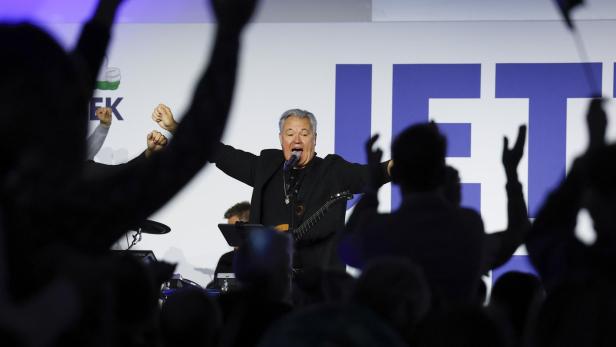 Ein Sänger mit Gitarre steht vor Publikum mit erhobenen Händen auf einer Bühne.