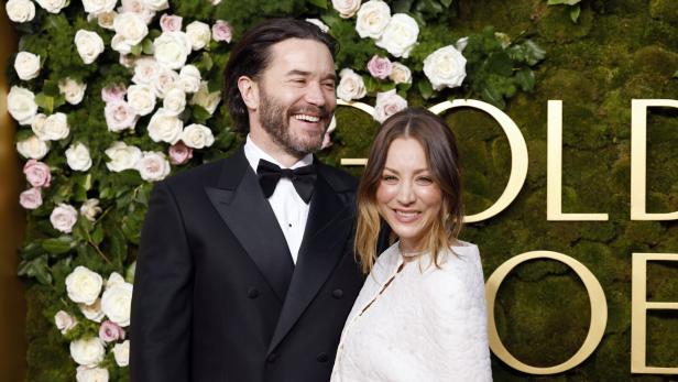 Kaley Cuoco und Tom Pelphrey, er im Smoking, sie in Weiß, posiert vor einem floralen Hintergrund.
