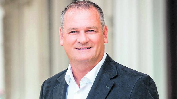 Ein Mann mit kurzem Haar und dunklem Sakko lächelt vor einem hellen Hintergrund.