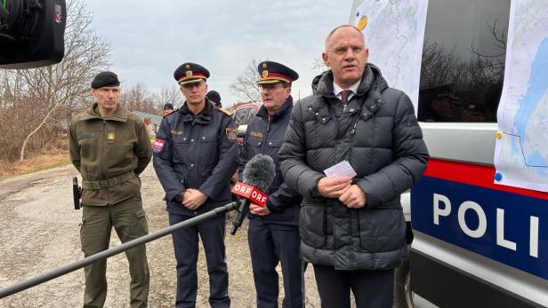Ein Mann in Zivilkleidung spricht vor Mikrofonen, flankiert von Polizisten vor einem Einsatzfahrzeug mit Karten.