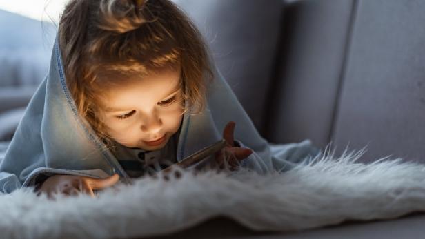 Ein kleines Mädchen liegt in eine Decke gehüllt und schaut auf ein leuchtendes Handy.