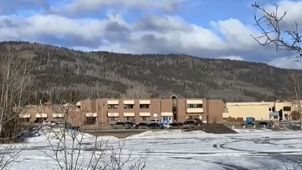 Ein langes, flaches Gebäude mit mehreren Autos davor steht vor einem schneebedeckten Gelände und bewaldeten Hügeln im Hintergrund.