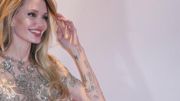 Angelina Jolie lächelt und berührt ihr Haar, ihr Arm mit Tattoos ist sichtbar.