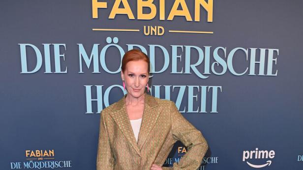 Eine Frau mit rotem Haar (Nicole Beutler) trägt einen gemusterten Hosenanzug und pinke Schuhe, steht vor einem blauen Hintergrund mit Schriftzug.