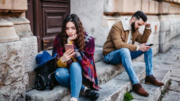 Eine Frau und ein Mann sitzen, voneinander abgewandt, auf Steinstufen, und starren auf ihre Smartphones.