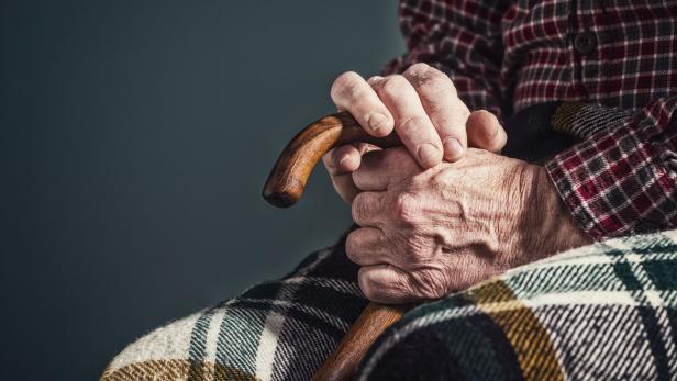Eine ältere Person hält einen Holzstock in ihren Händen, bedeckt mit einer karierten Decke.
