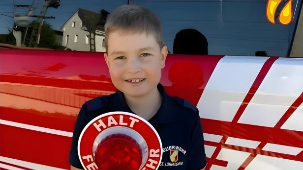 Ein Junge in Feuerwehrkleidung hält ein Stoppschild vor einem roten Feuerwehrfahrzeug.