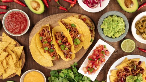 Mexikanisches Essen mit Tacos, Tortilla-Chips, Guacamole, Salsa, Käse, Limetten, Chili und Jalapeños auf Holztisch.