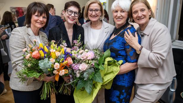 Fünf Frauen stehen eng beisammen, zwei von ihnen halten große bunte Blumensträuße und alle lächeln.