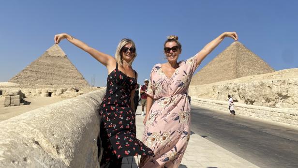 Zwei Frauen posieren vor den Pyramiden von Gizeh mit erhobenen Armen.