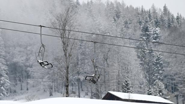 Zwei leere Sessellifte hängen vor einem verschneiten Wald und einem kleinen Haus.