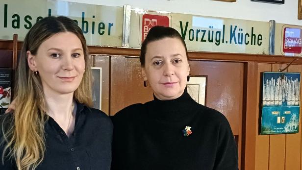 Zwei Frauen stehen nebeneinander in einem Raum mit Holzvertäfelung und alten Werbeschildern an der Wand.