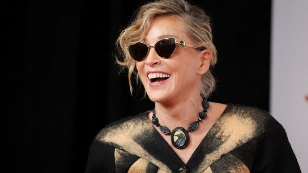 Frau mit Sonnenbrille und auffälliger Halskette lacht, trägt ein schwarz-gelbes Oberteil vor dunklem Hintergrund.