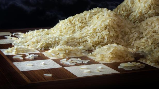Reis liegt in mehreren Haufen und einzelnen Körnern auf einem Schachbrett mit dunklem Hintergrund.