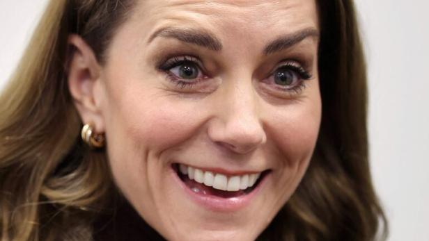 Prinzessin Kate mit braunen Haaren und braunen Augen lächelt freundlich, trägt braunen Rollkragenpullover und Ohrringe.