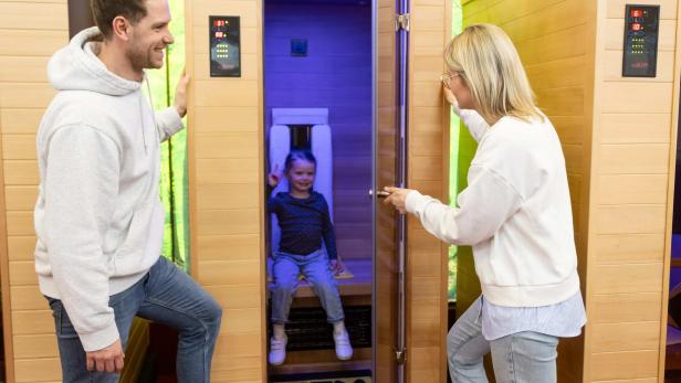 Ein Kind sitzt in einer Infrarotkabine, zwei Erwachsene stehen lächelnd davor.