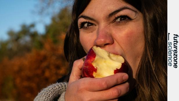 Frau beißt in einen roten Apfel im Freien mit herbstlichem Hintergrund.
