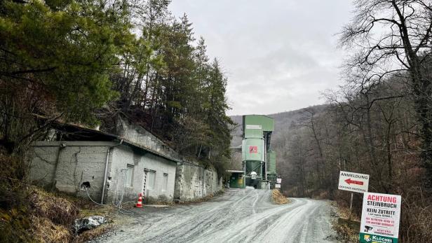 Eine Schotterstraße führt zu einer Industrieanlage im Wald, mit Warnschildern und einem alten Gebäude am Straßenrand.