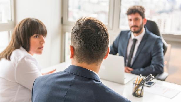 Zwei Männer und eine Frau sitzen in Business-Klamotten in einem lichtdurchfluteten Büroraum. 