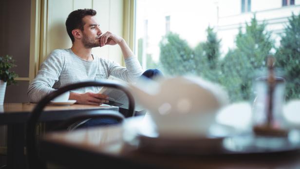 Mann sitzt nachdenklich mit Zeitung an einem Tisch am Fenster in einem Café.