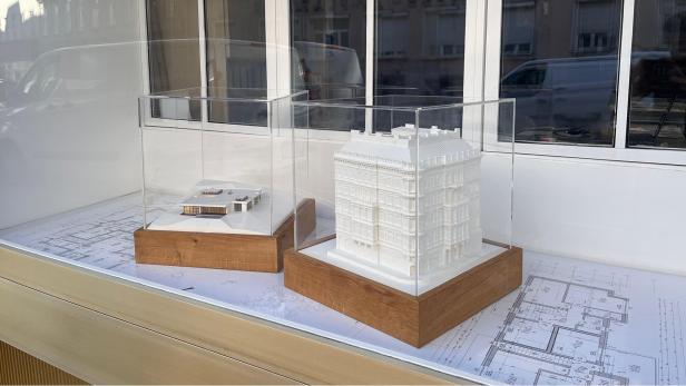 Zwei Architekturmodelle in Vitrinen auf Holzsockeln, ausgestellt auf Bauplänen vor einem Fenster.