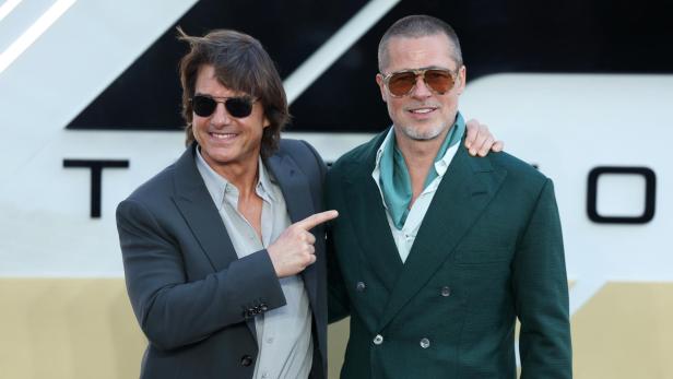 Tom Cruise zeigt auf Brad Pitt, beide tragen Anzüge und Sonnenbrillen.