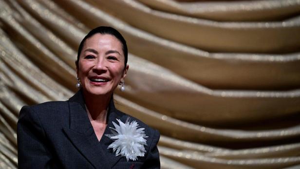 Eine Frau mit dunklem Haar - Michelle Yeoh - trägt einen dunklen Blazer mit großer weißer Blume und Perlenohrringen vor goldenem, gerafftem Hintergrund.