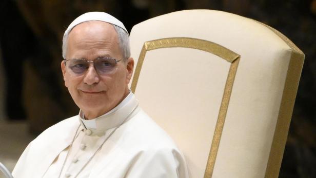 Ein Mann mit Brille und weißem Gewand sitzt auf einem verzierten Stuhl.