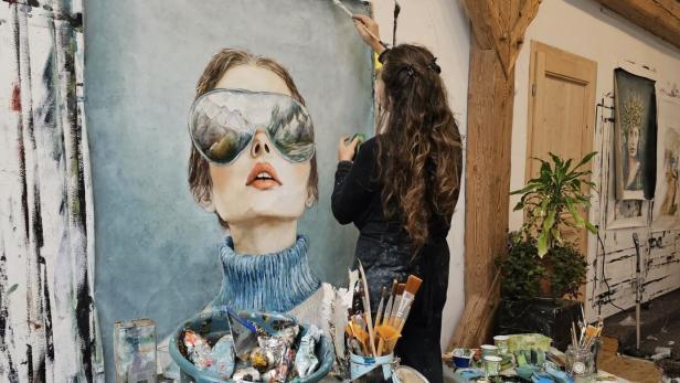 Doris Breuer arbeitet an einem ihrer Gemälde an der weißen Stadlwand. Zu sehen ist eine Frau in blau-tönen mit einer Skibrille, in der sich die Berge spiegeln.
