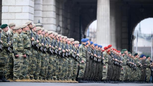 Mehrere Reihen Soldaten in Uniform und mit verschiedenfarbigen Baretten stehen in Formation vor einem Gebäude mit Säulen.