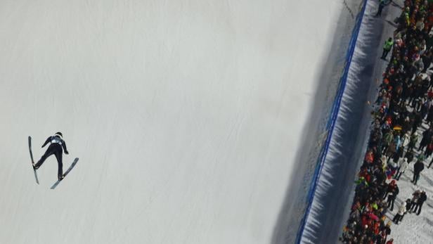 Ein Skispringer ist in der Luft über einer verschneiten Piste, während Zuschauer am Rand stehen.