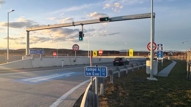 Eine Autobahnauffahrt mit Verkehrszeichen, Ampeln und einem Auto ist zu sehen.