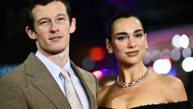 Callum Turner und seine Partnerin, Sängerin Dua Lipa mit Halskette und schulterfreiem Kleid, stehen nebeneinander vor bunten Lichtern.