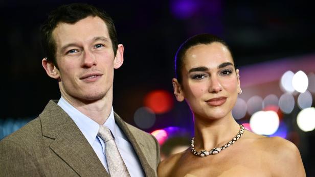 Callum Turner und seine Partnerin, Sängerin Dua Lipa mit Halskette und schulterfreiem Kleid, stehen nebeneinander vor bunten Lichtern.