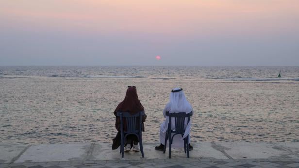 Eine Frau mit schwarzem Umhang und ein Mann mit weißer Kleidung schauen in den Sonnenuntergang