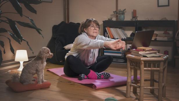 Eine Frau macht Dehnübungen auf einer Yogamatte vor einem Laptop, während ein kleiner Hund auf einem Kissen sitzt.