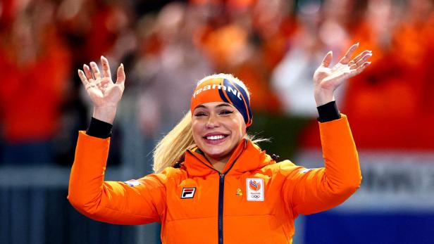 Eine Frau in oranger Sportkleidung lächelt und hebt beide Hände jubelnd in die Luft, im Hintergrund jubelnde Menschen.