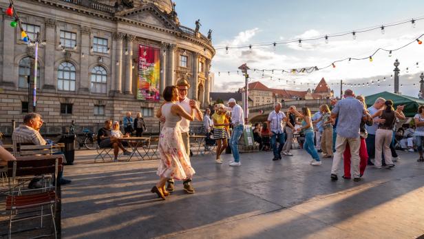 Mehrere Menschen tanzen fröhlich im Freien vor einem historischen Gebäude bei Sonnenuntergang.