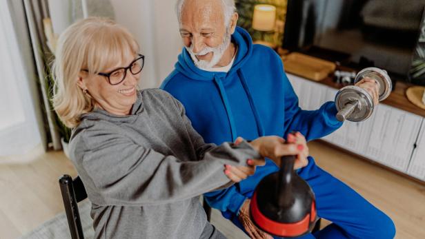 Ein älteres Paar trainiert gemeinsam mit Hantel und Kettlebell und lächelt dabei.