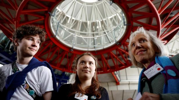 Drei Menschen stehen unter einer großen, roten, kreisförmigen Stahlkonstruktion und blicken in verschiedene Richtungen.