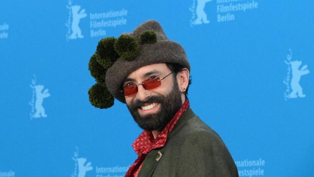 Eine Person mit auffälligem Hut, rotem Hemd, grünem Sakko und Sonnenbrille posiert vor blauem Berlinale-Hintergrund.