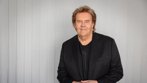 Howard Carpendale mit blondem Haar und schwarzem Sakko lächelt vor einem hellen Hintergrund.