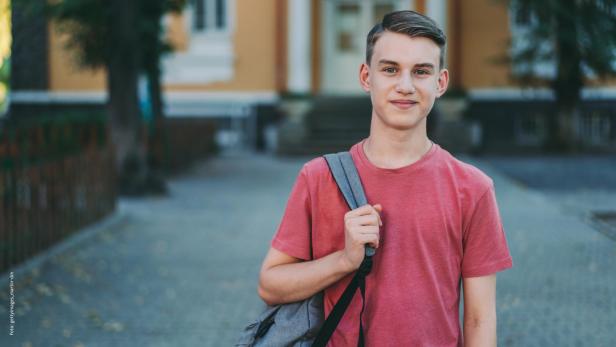 Jugendlicher mit Rucksack steht draußen vor einem Gebäude und lächelt in die Kamera.