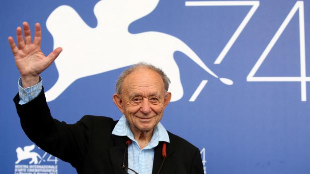 Ein älterer Mann im Anzug - Frederick Wiseman - winkt vor einer blauen Wand mit weißem Löwen-Logo und der Zahl 74.
