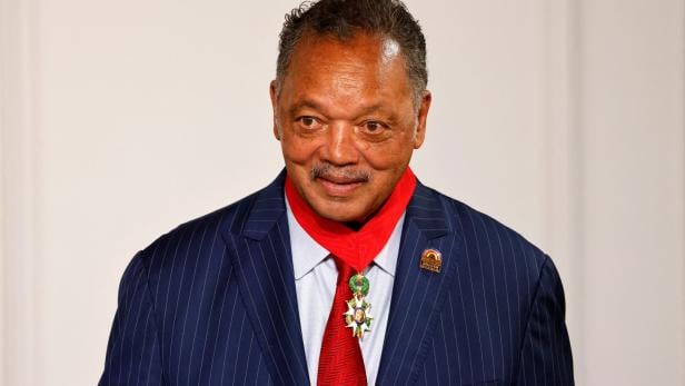 Jesse Jackson trägt einen Anzug mit Nadelstreifen, eine rote Krawatte und eine Auszeichnung.
