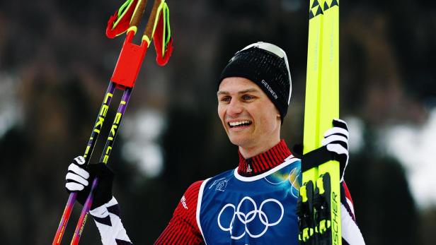 Ein lächelnder Skilangläufer mit Mütze hält Skier und Stöcke in der Hand.