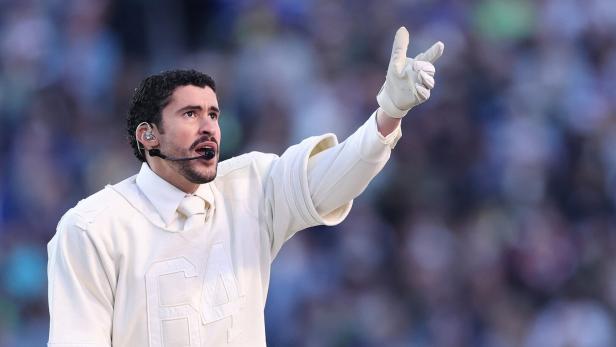 Musiker Bad Bunny mit Headset und weißen Handschuhen trägt ein weißes Oberteil mit der Zahl 64 und zeigt nach oben.