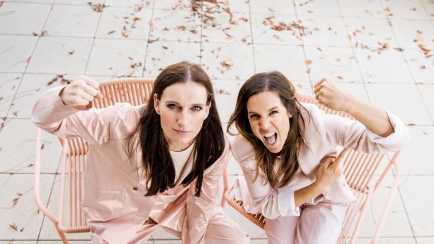 Zwei Frauen in rosa Anzügen sitzen auf Stühlen, ballen die Fäuste und schauen entschlossen nach oben.